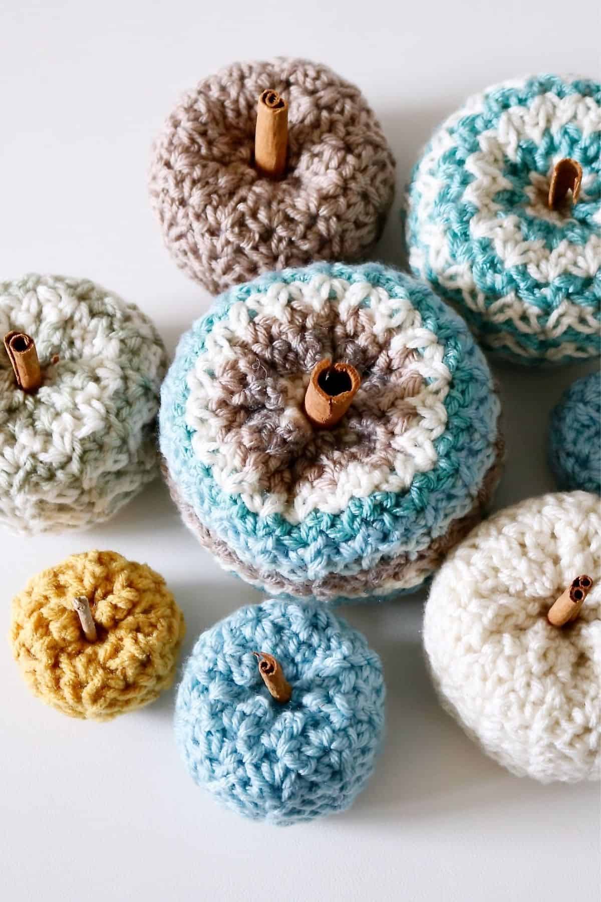 Pastel-colored crocheted pumpkins with cinnamon stick stems are arranged on a white surface, displaying the Cedar Ridge Pumpkins free crochet pattern. Free crochet pattern