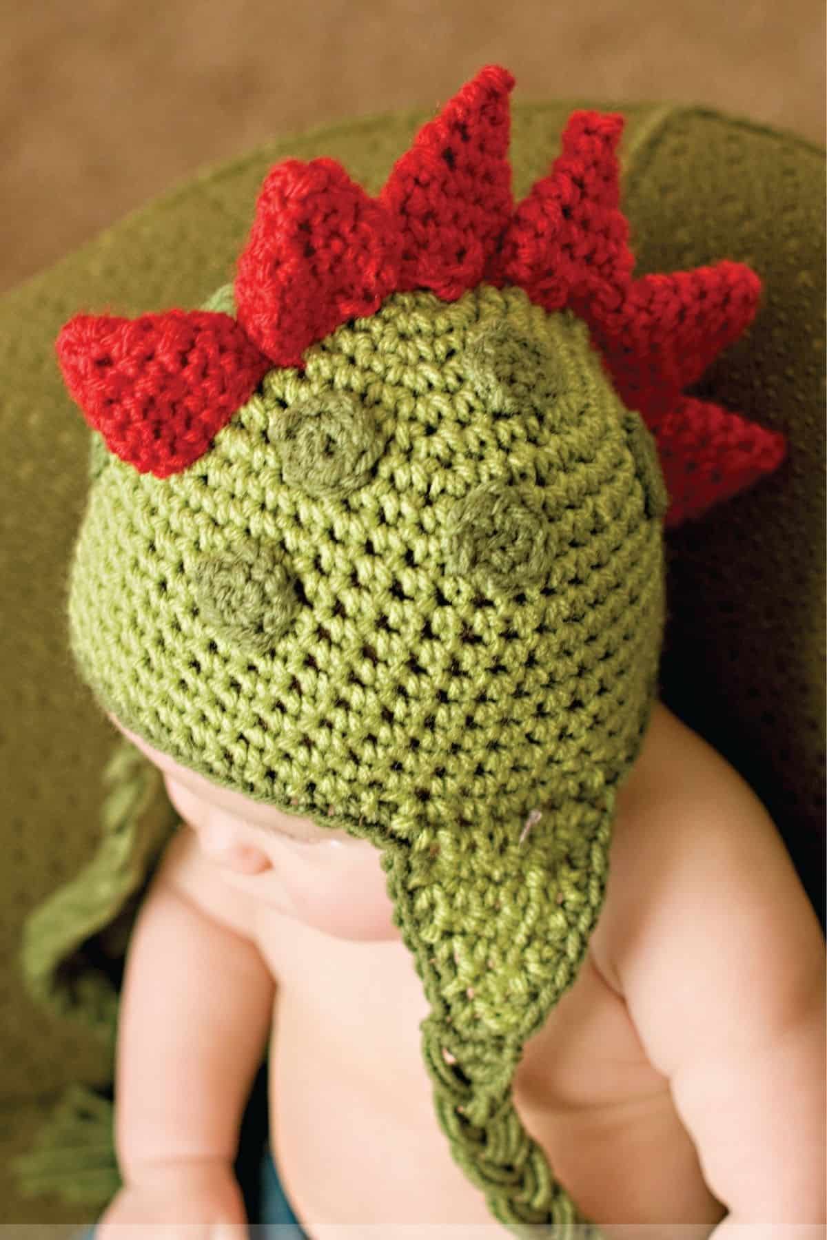 A baby wearing a green dinosaur hat with red spikes along the center. Free crochet pattern